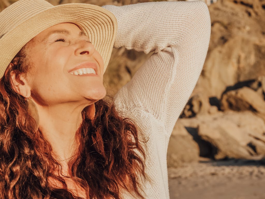 A joyful woman with a hat basks in the sunlight by the seaside, symbolizing relaxation and happiness.